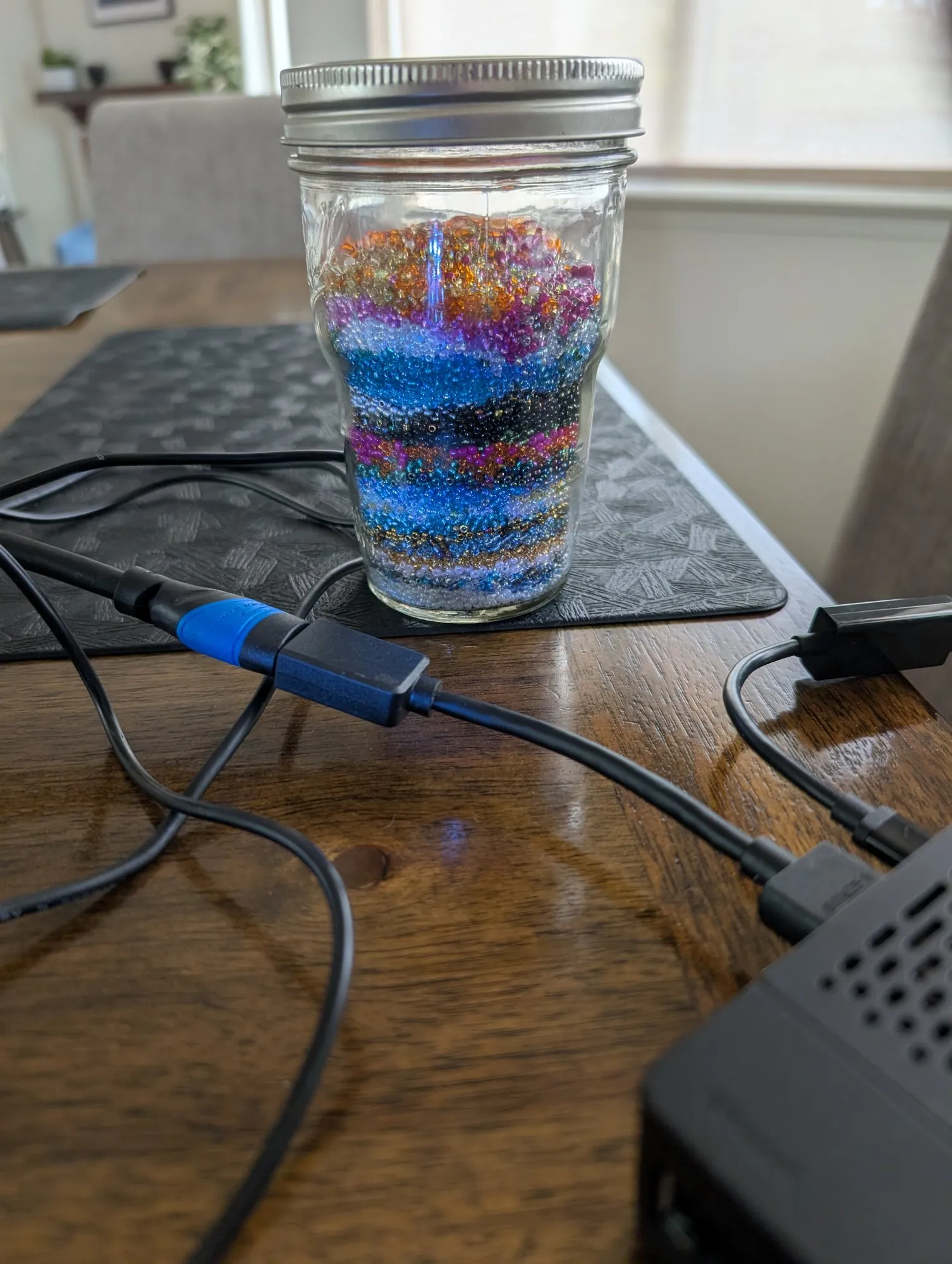A mason jar of layered seed beads in bands of color on a table, cables around it.