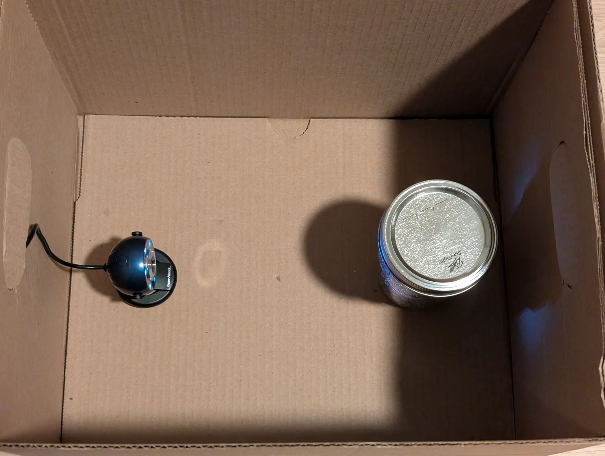 An open cardboard box seen from above: a mason jar with a Ball lid stands at the right, and a webcam at the left points at it, a ring of LEDs lit around its lens and its cable running out a handle hole.