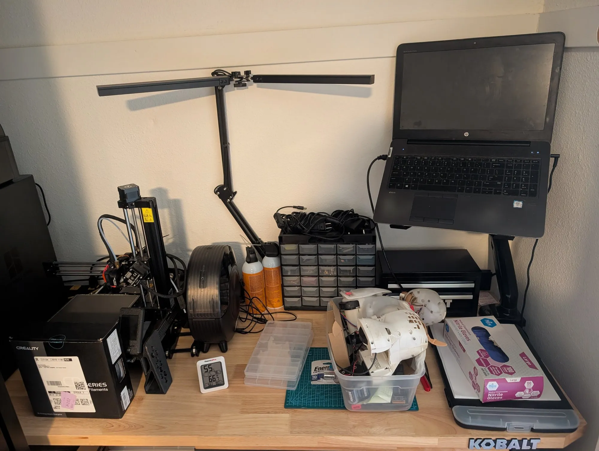 The electronics bench: the Prusa Mini at the left beside a black filament spool, a desk lamp, a small parts organizer, and a bin holding the gutted white shell of Bubbles the robot dog, with the HP ZBook on an arm mount at the right.