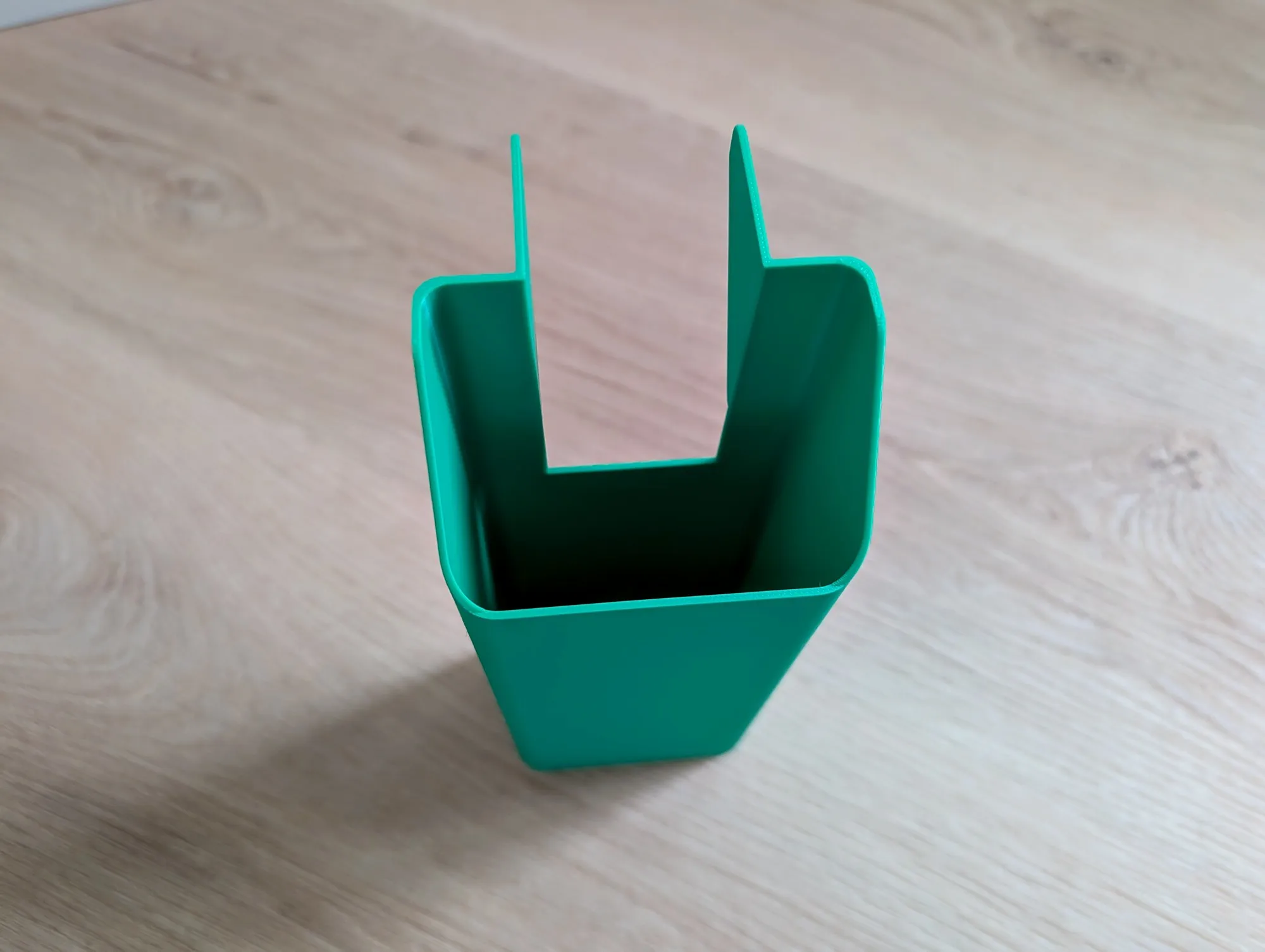 A green 3D printed chute, open on top with a U-shaped slot, standing on a wood floor.