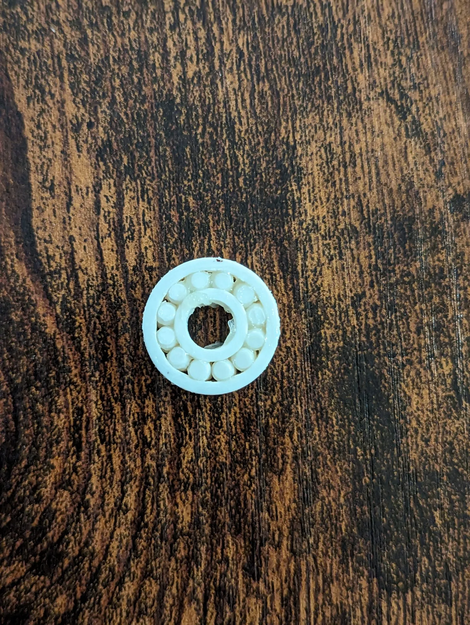 A tiny white 3D printed ball bearing, complete with printed balls in the race, sitting on a dark wood table.