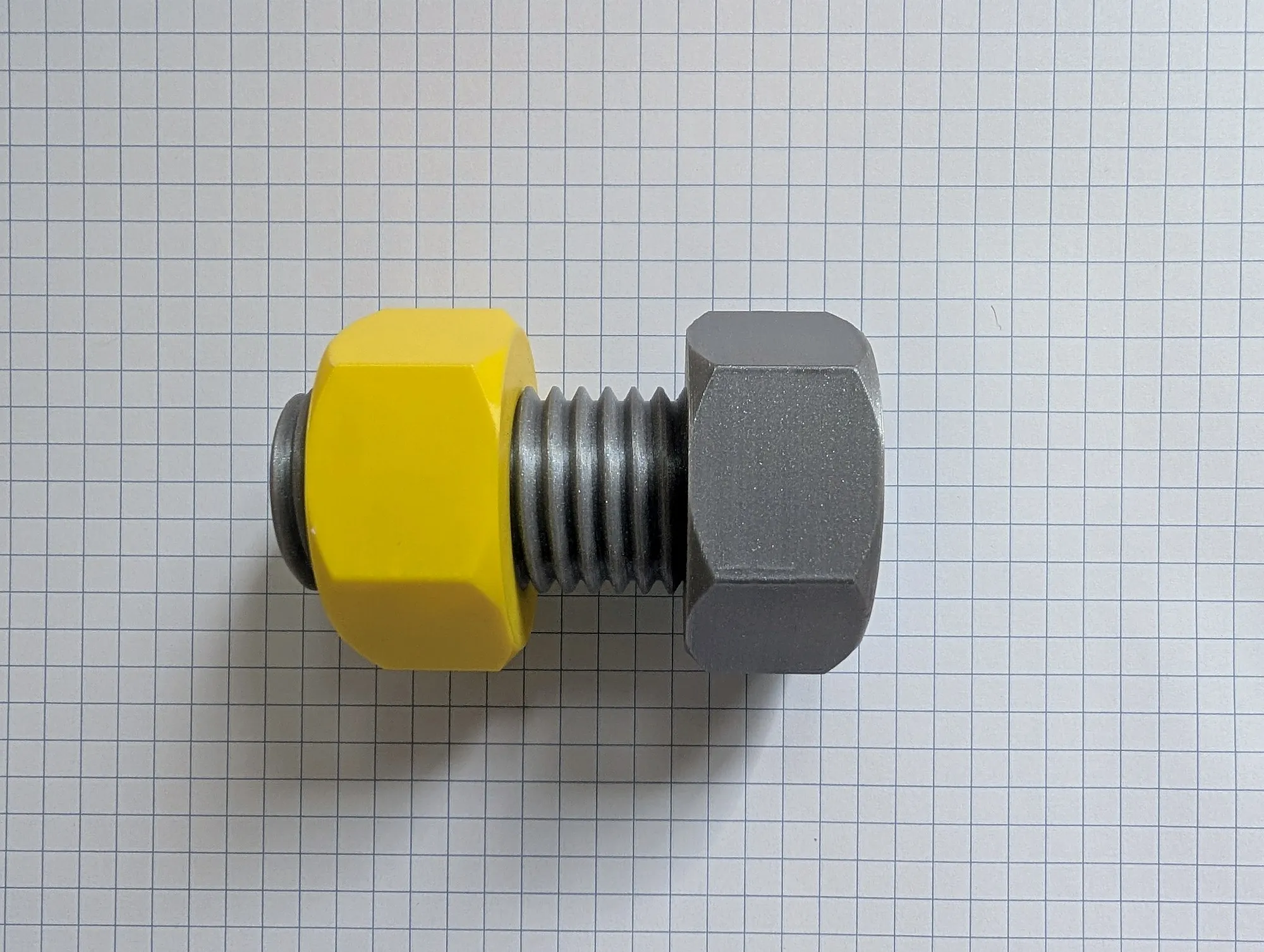 An oversized 3D printed bolt in silver gray threaded into a yellow hex nut, lying on grid paper.