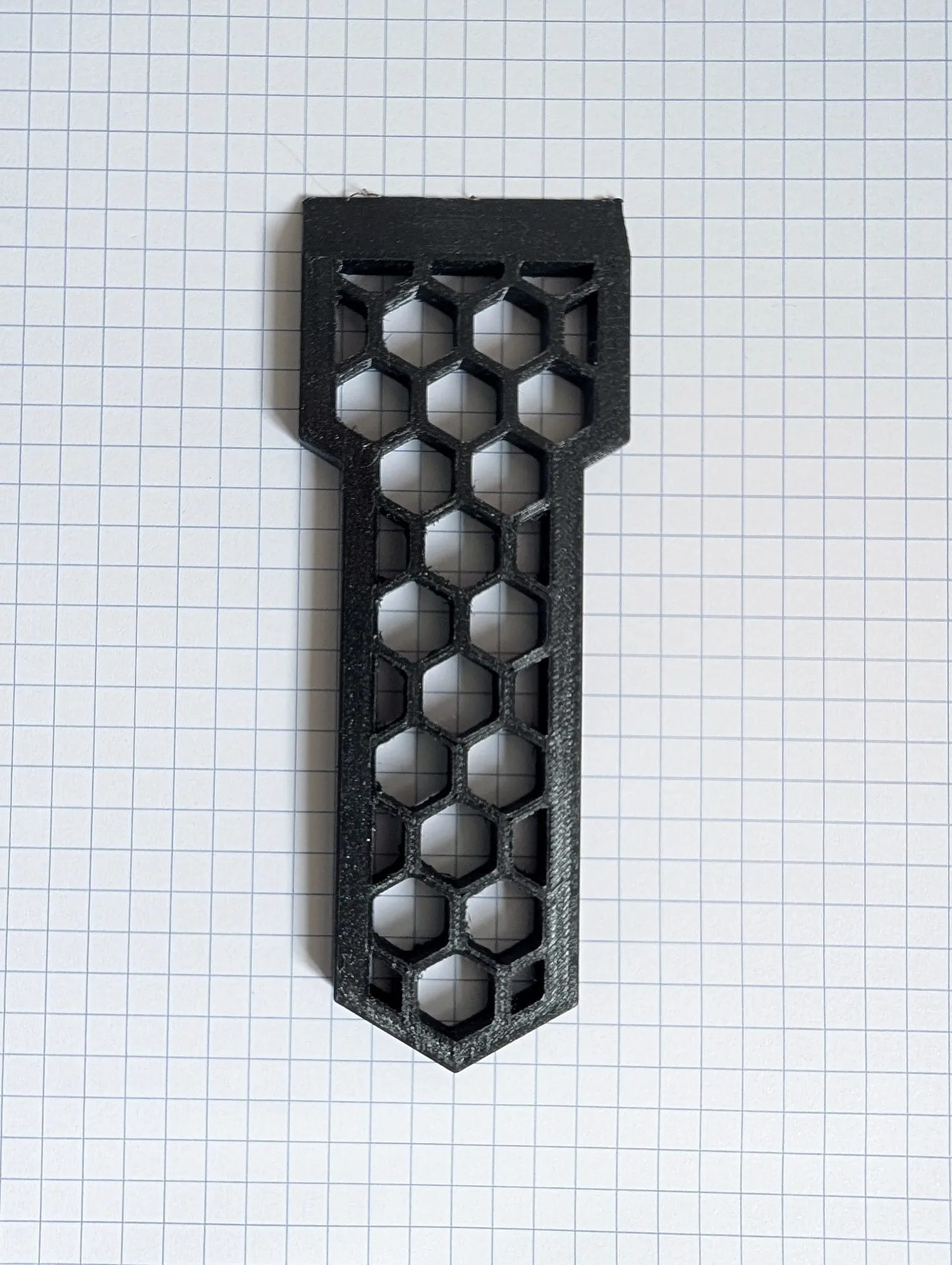 A black 3D printed parts scraper with a honeycomb cutout handle, lying on grid paper.