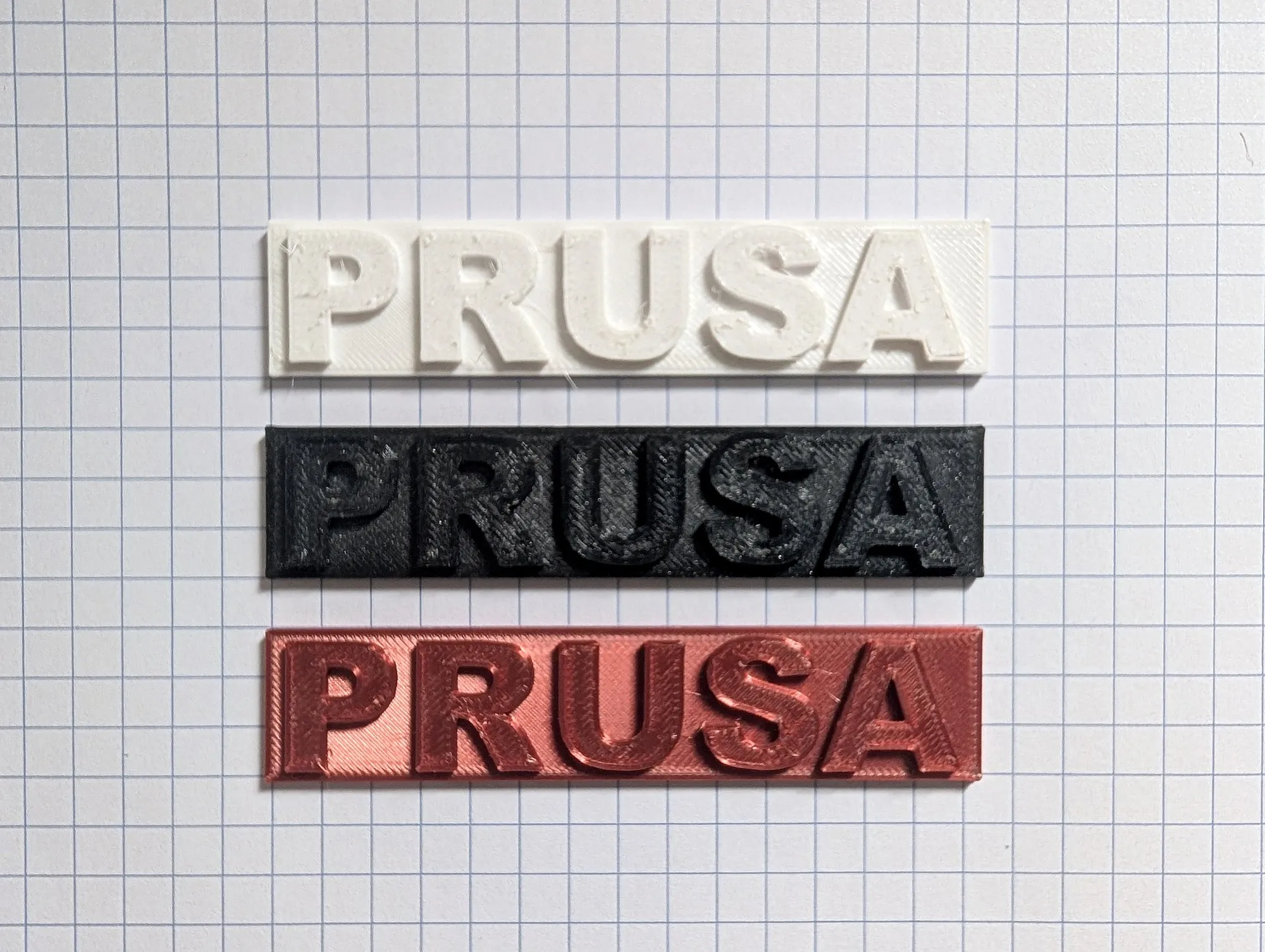 Three 3D printed PRUSA logo plates stacked on grid paper, one white, one black, and one copper.