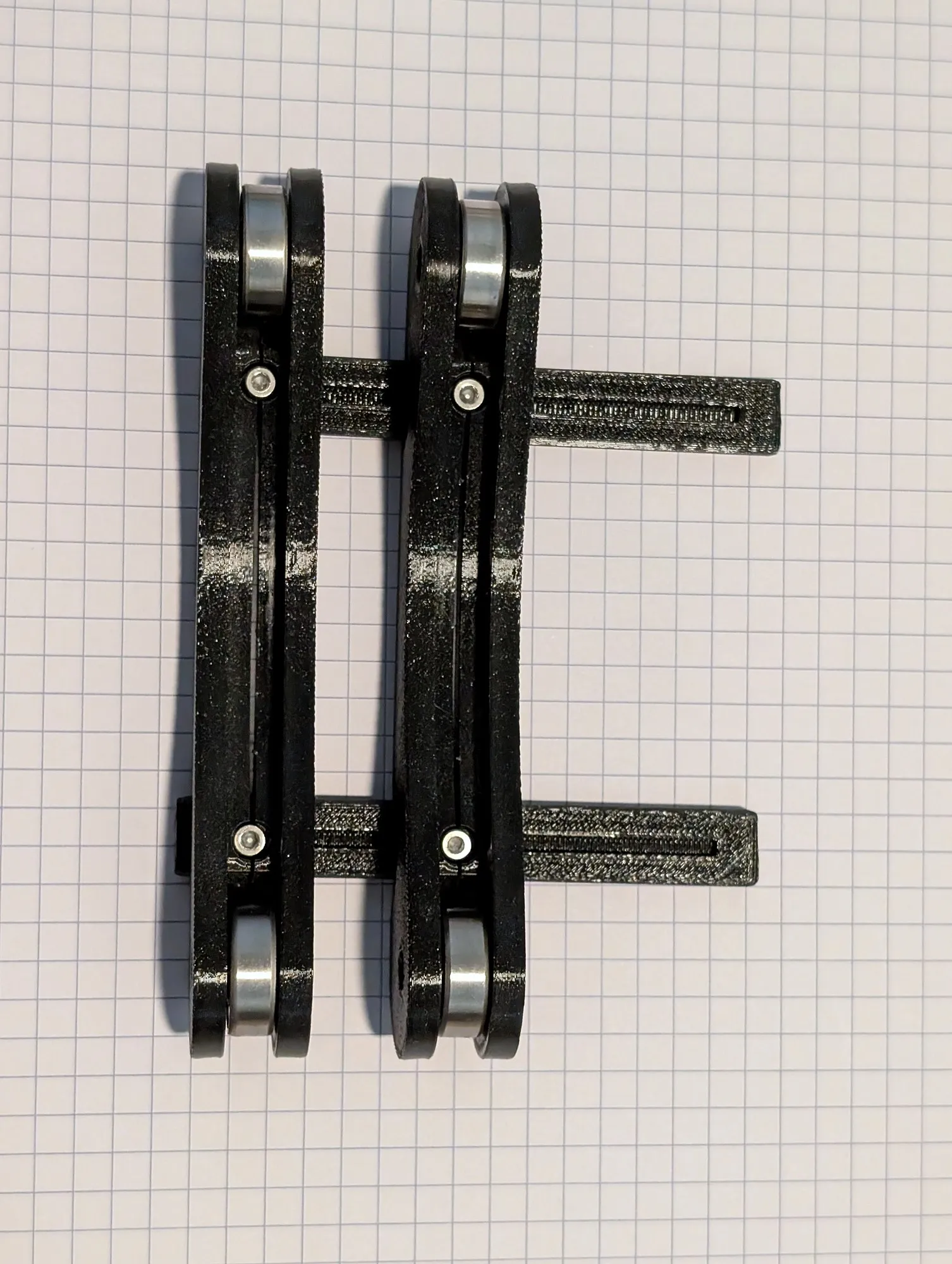 A black 3D printed adjustable spool holder on grid paper, two long arms each holding a steel bearing at both ends, joined by two slotted bars.
