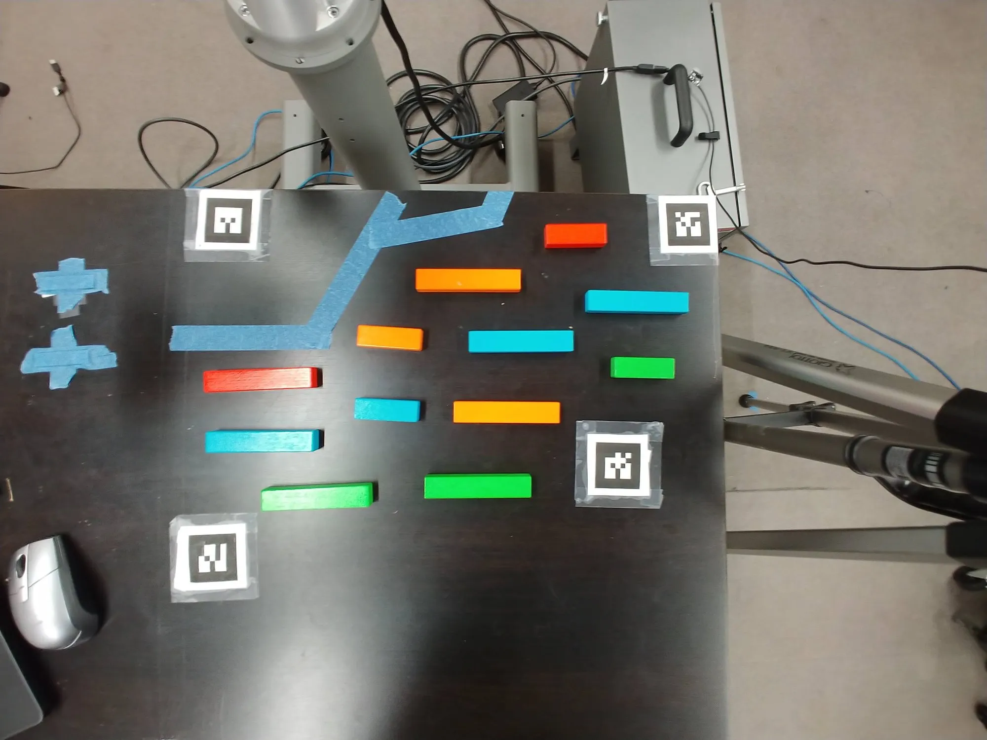 Blocks of four colors scattered on the black table between strips of blue tape, ArUco tags taped at the corners, the arm's base at the top edge.