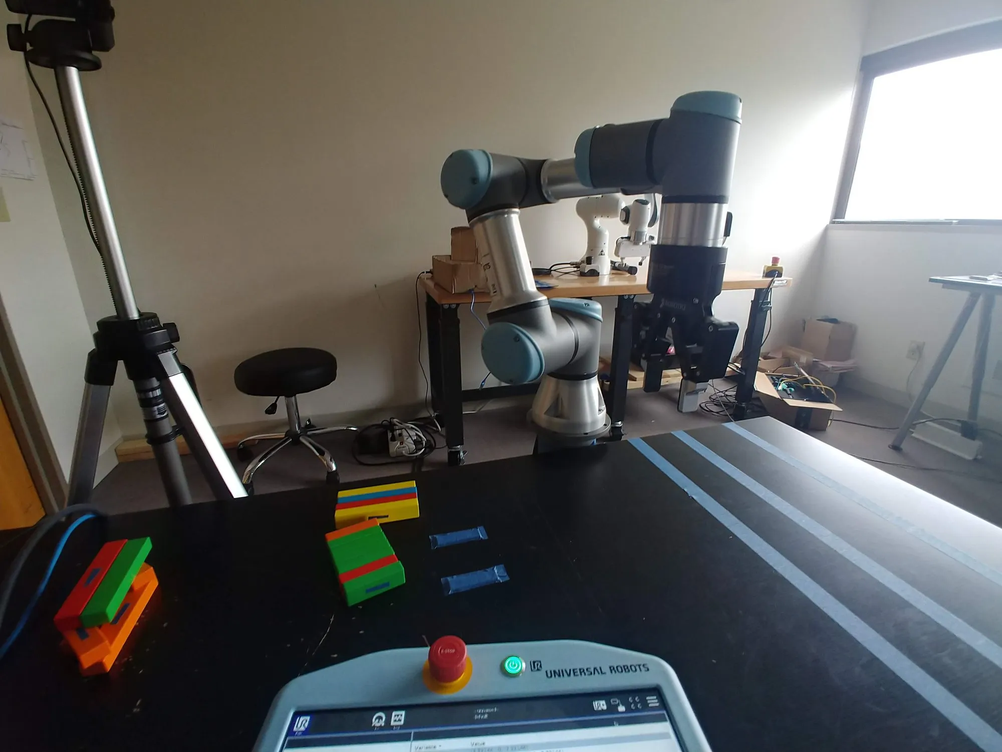 The setup: the UR3e on its bench with a tripod camera beside it, colored blocks on the black table in front, the teach pendant at the near edge.