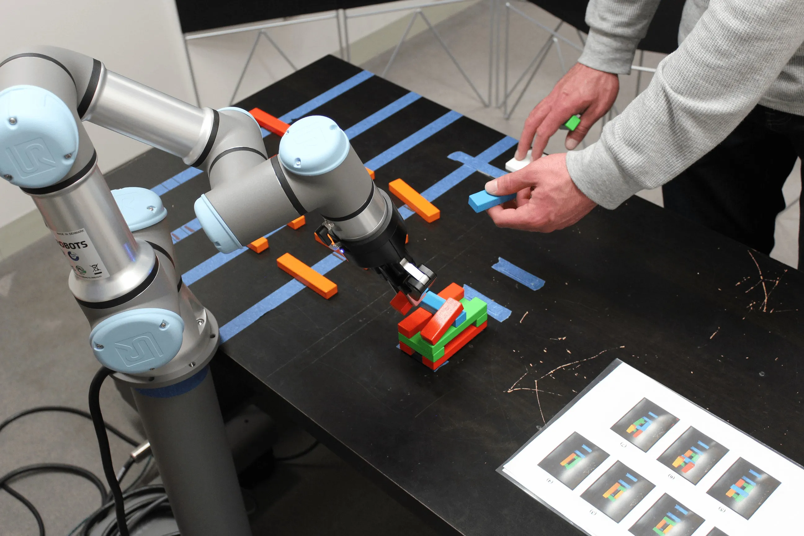 A participant's hands and the UR3e arm building block structures together on a taped-off table, with a sheet of target patterns at the edge