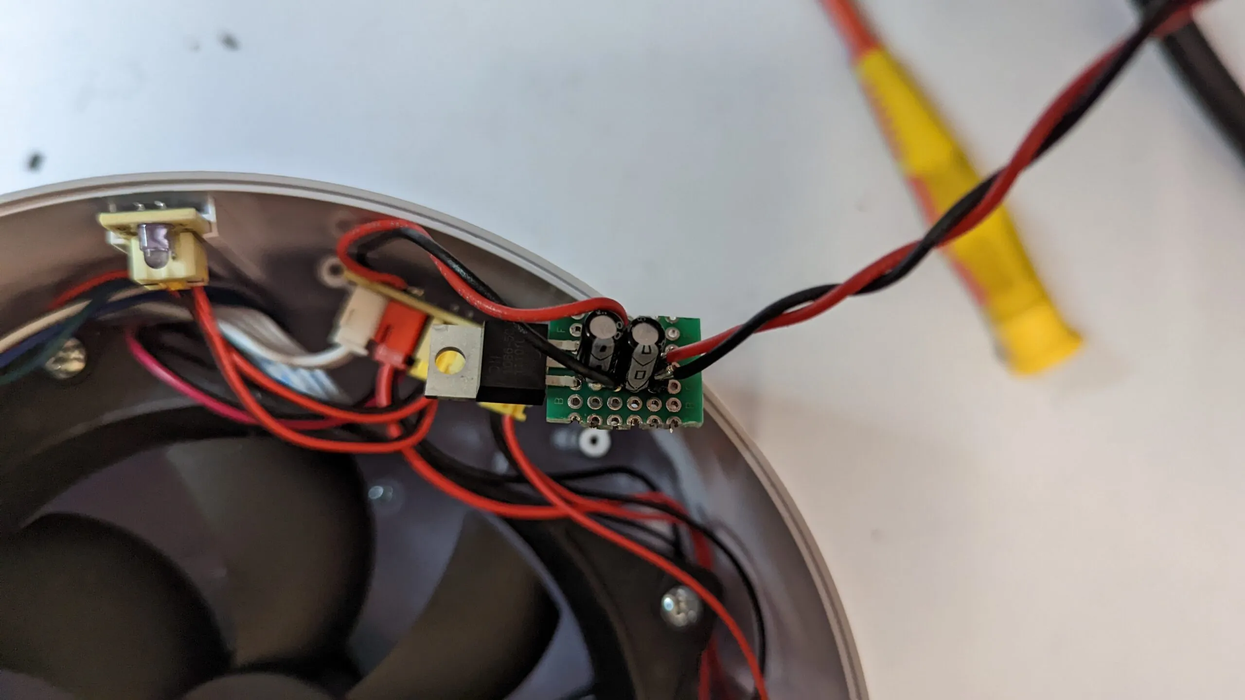 The step-down regulator on a scrap of perfboard, a TO-220 regulator and two capacitors, twisted red and black leads, sitting on the rim next to the fan