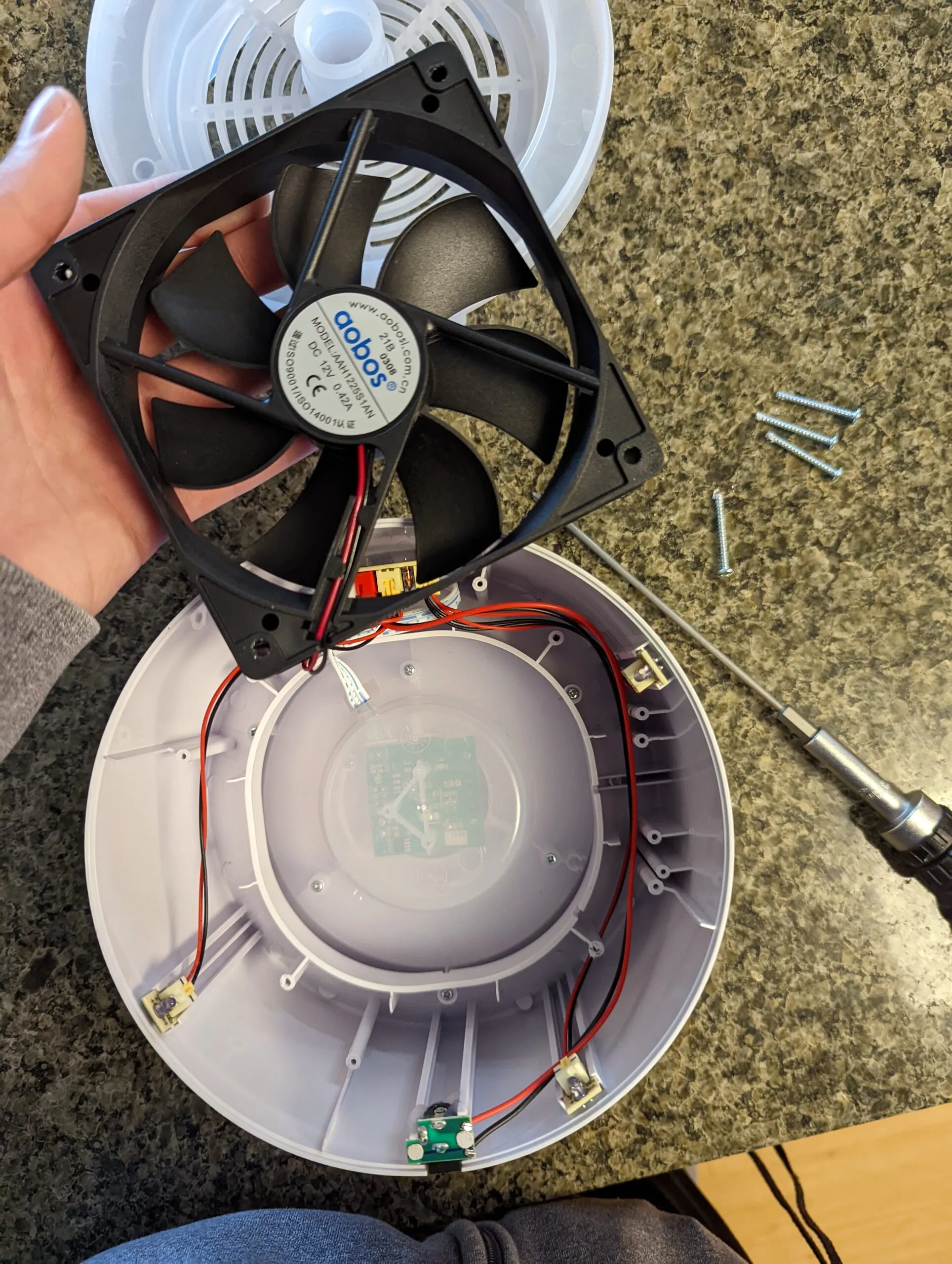 The 12 V Aobos fan freed from the housing, mounting screws scattered on the counter, the control board showing through the translucent center of the shell