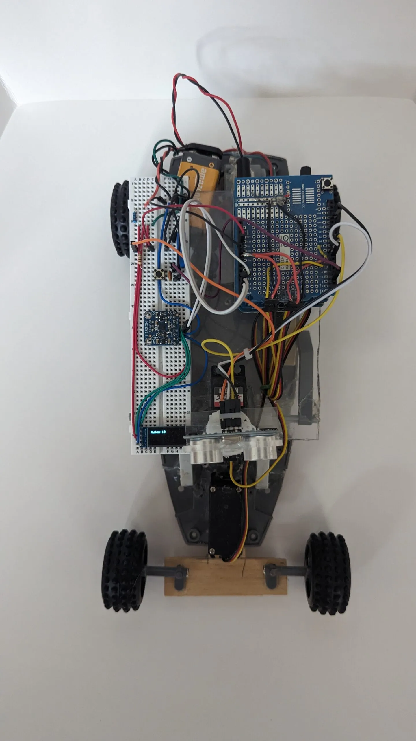 Top-down view of RoverBot: breadboard deck, 9V, and shield stack over the gray chassis, wooden axle at the front