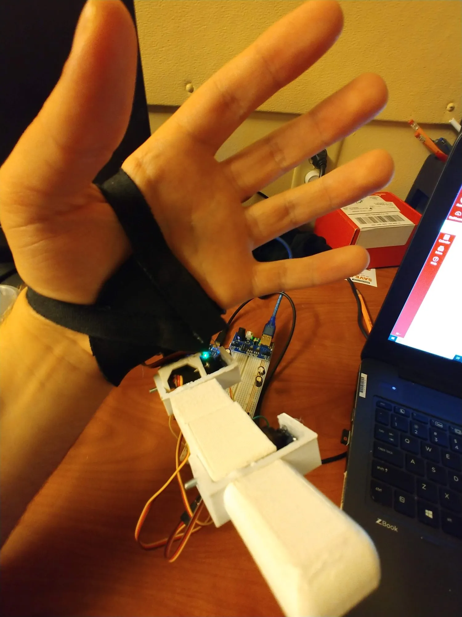 The first printed finger on the brace, angled out beside the pinky, dev board on the desk behind.