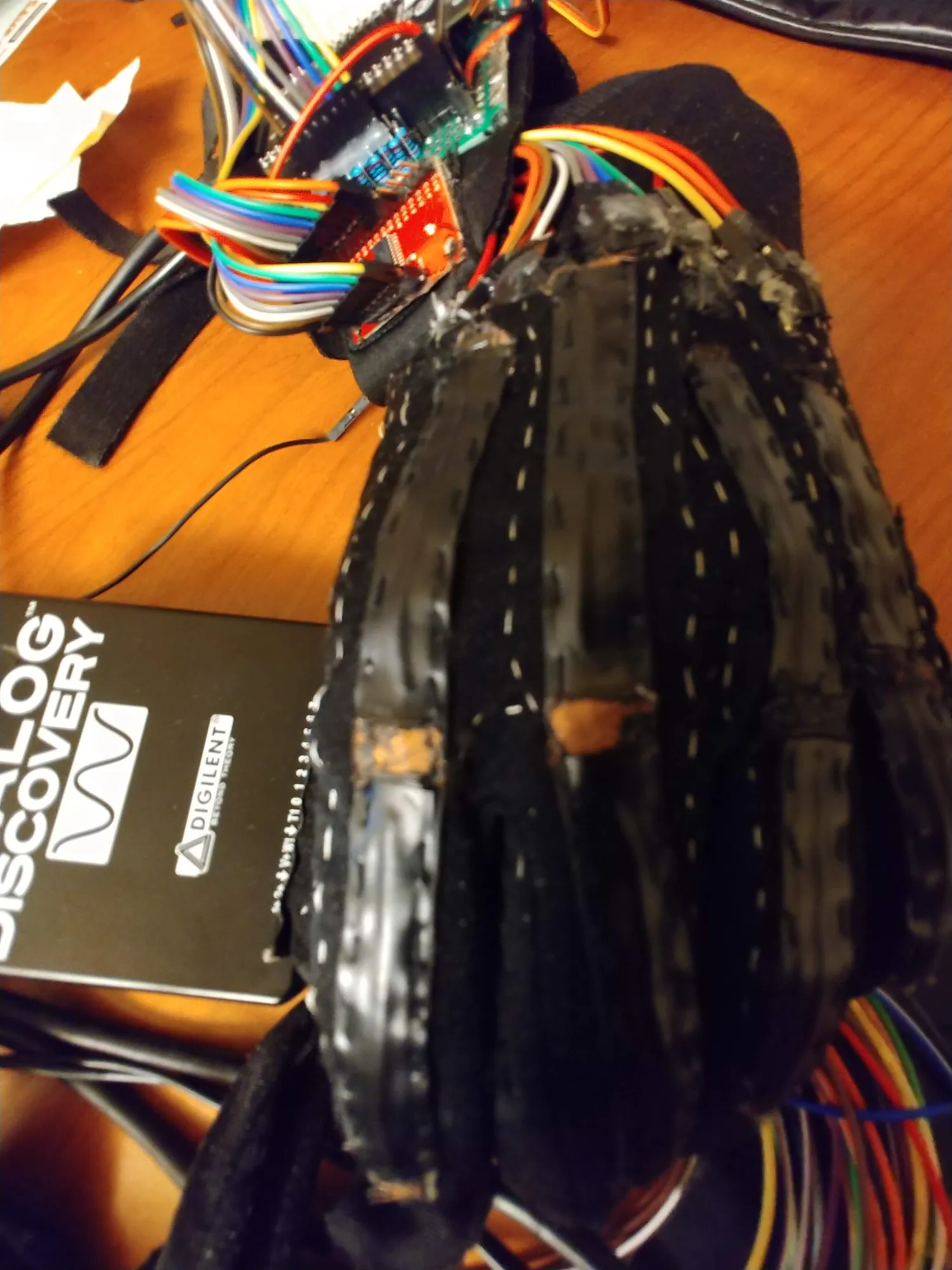 The black glove with taped conductive strips down each finger and a rainbow ribbon of leads at the wrist.