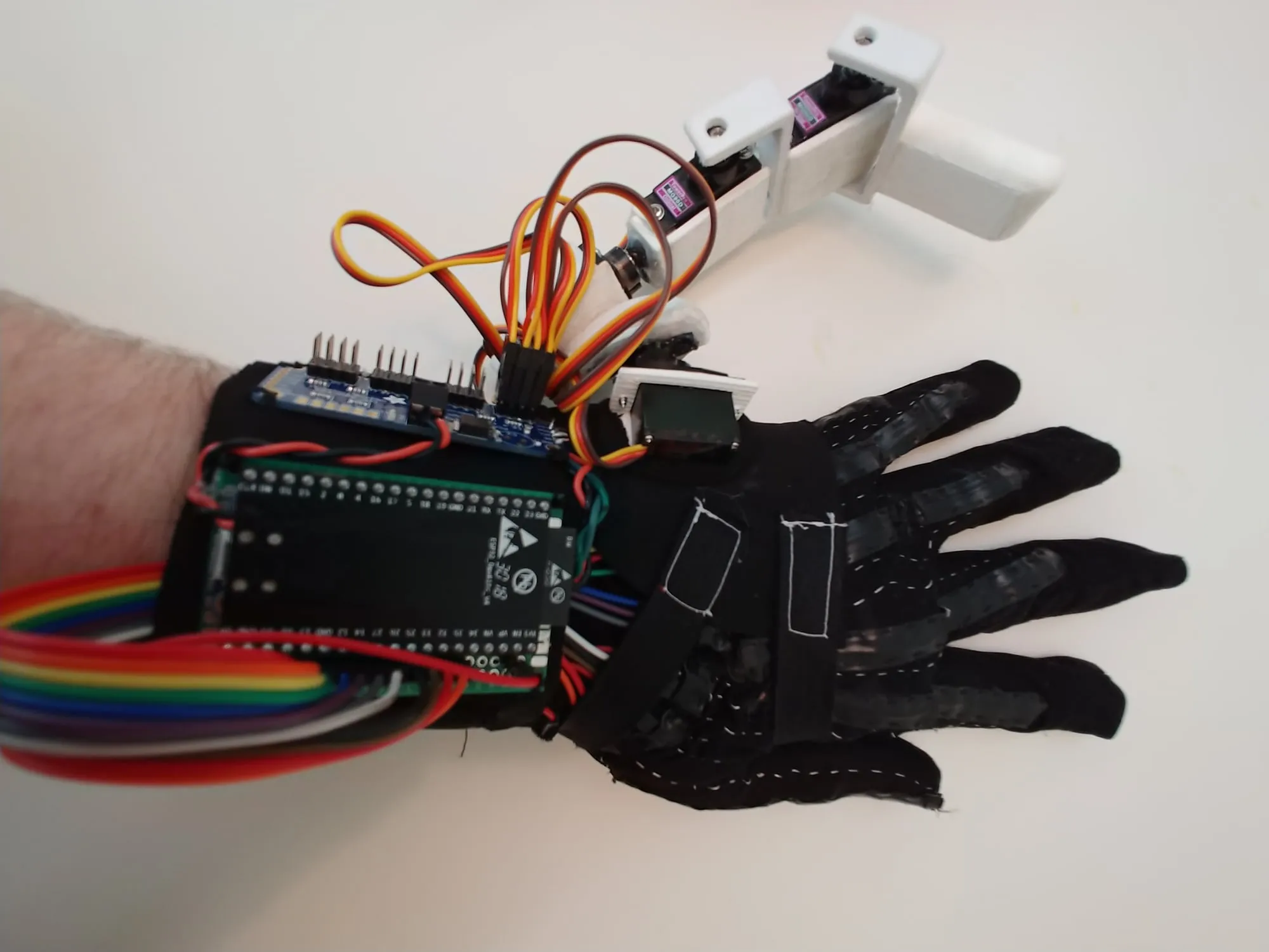 Top-down view of a black glove with home-made flex sensors sewn along each finger, an ESP32 dev board and servo driver strapped to the wrist with rainbow ribbon cable, and the 3D printed robotic finger with its servos and orange servo leads mounted beside the little finger.