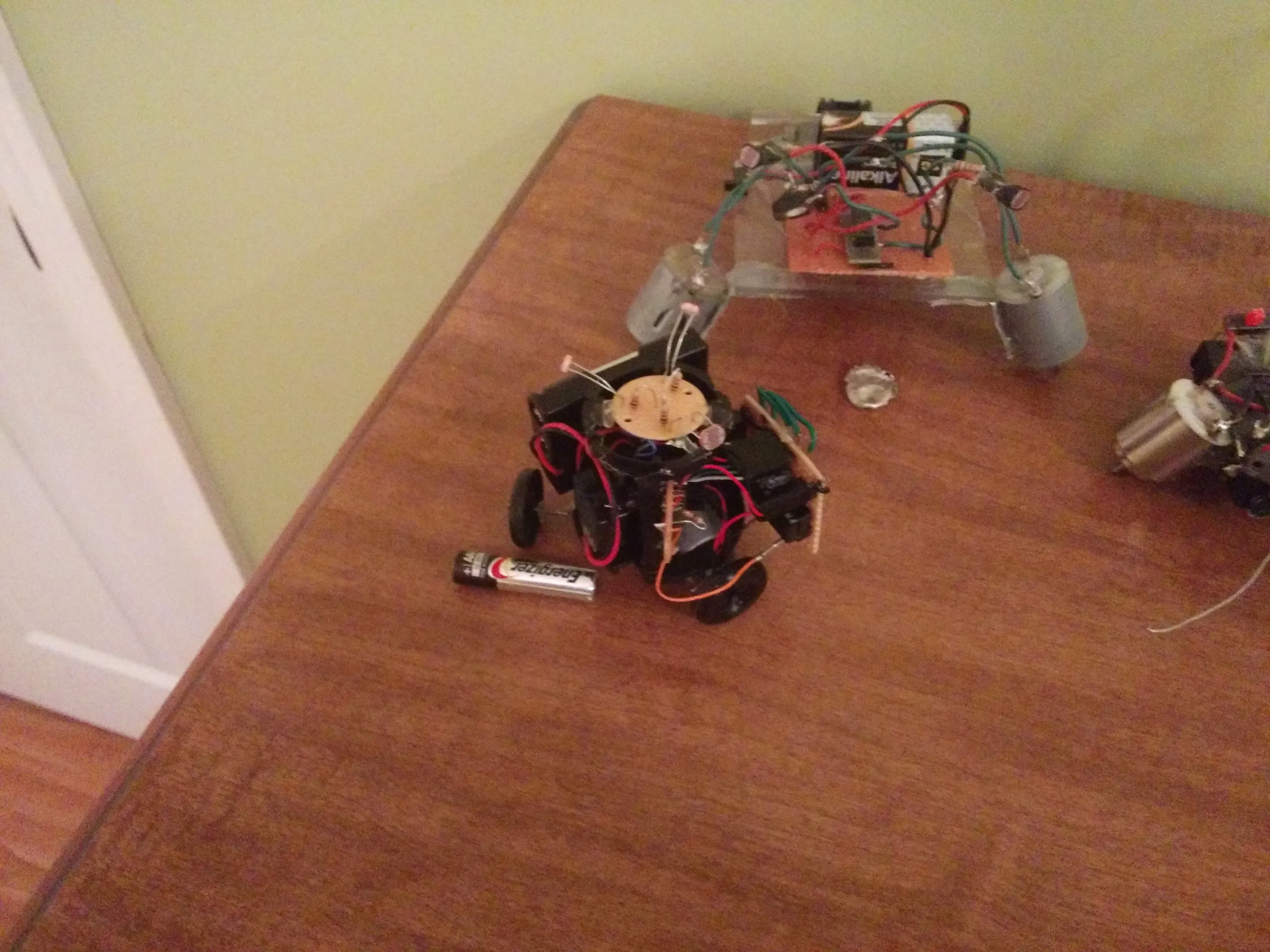 Triclops posed beside an acrylic-framed sibling robot, spare gearmotor at the edge of frame
